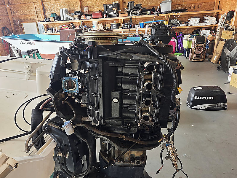 An outboard powerhead stripped down on the stand in the shop, a Suzuki cowl on the floor